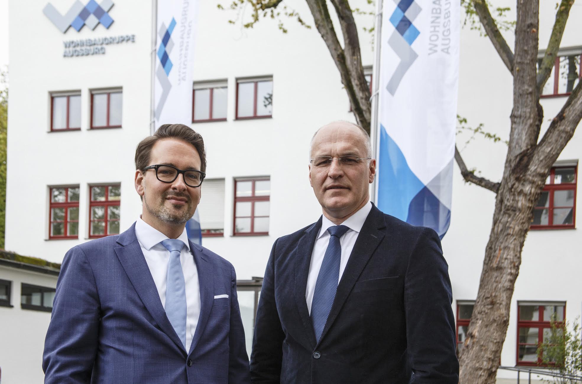 Mit neuem Logo im Hintergrund: Dr. Mark Dominik Hoppe (li.), und Oberbürgermeister Dr. Kurt Gribl vor der Geschäftsstelle der Wohnbaugruppe Augsburg.